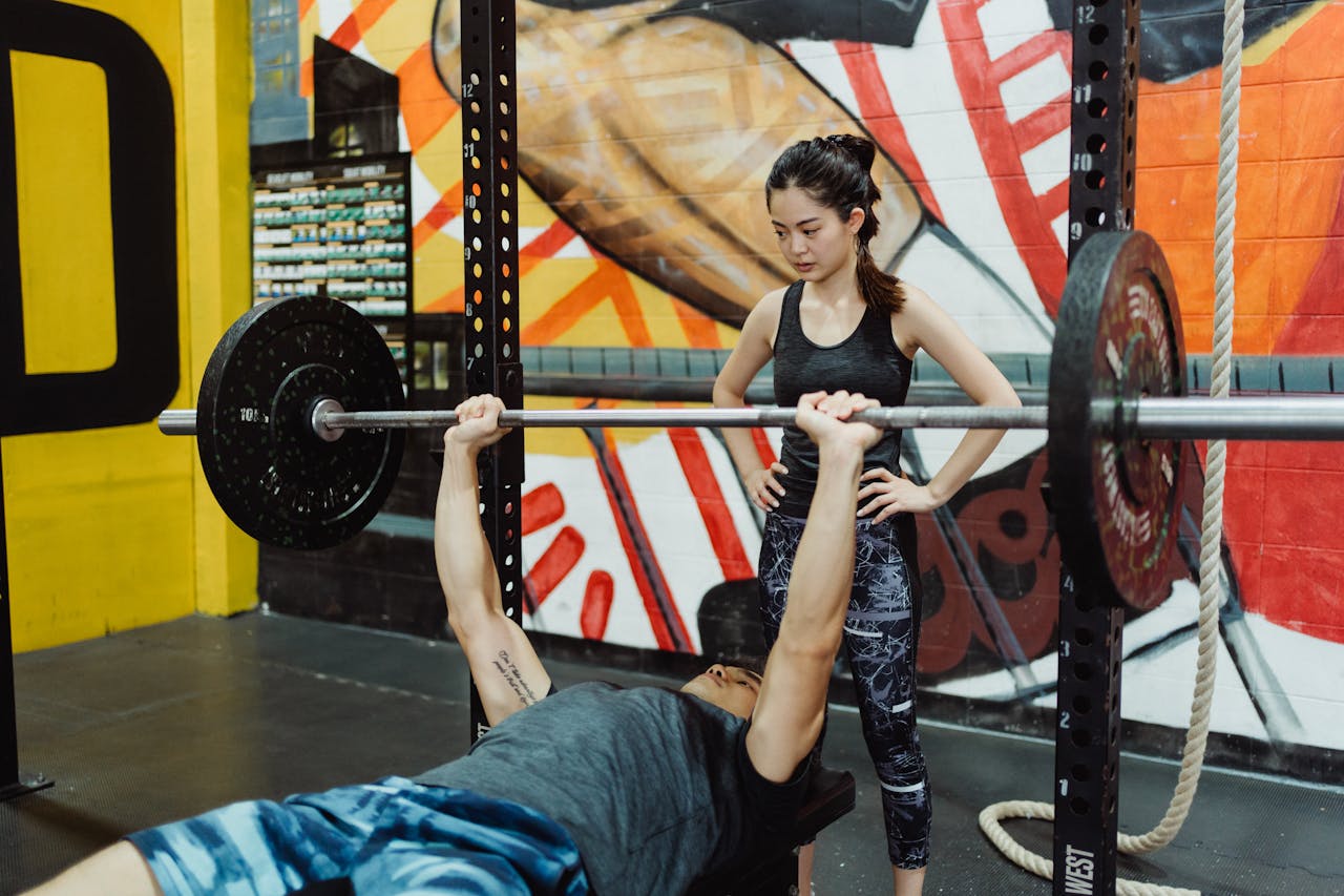 A man bench pressing in a gym with a mural and a woman spotting him.